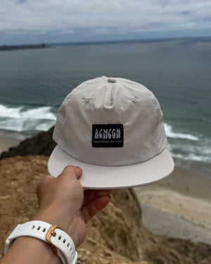 Beige cap with a brand logo held by a hand on a beach with ocean and sky in the background